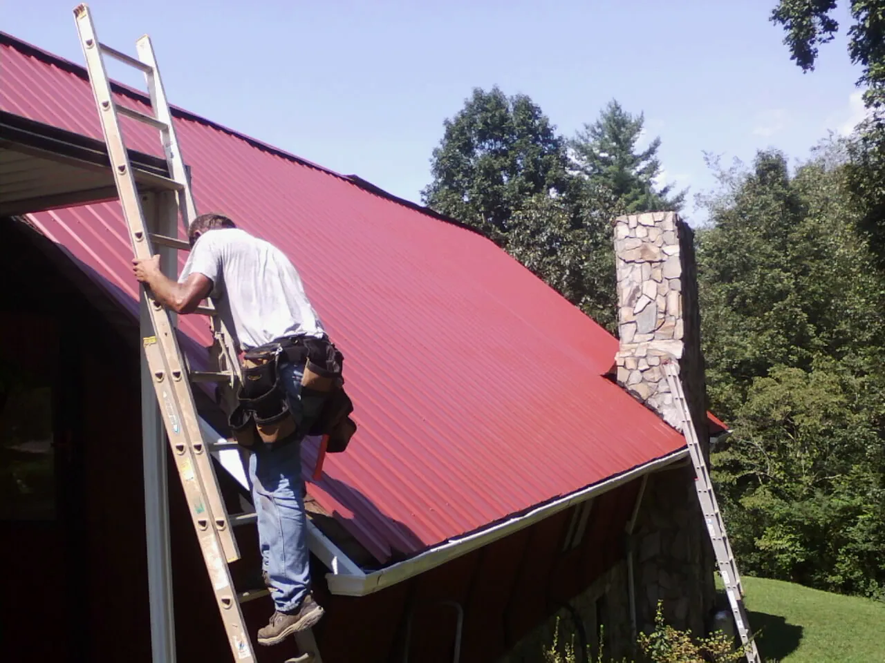 Roofer on a ladder beside a red metal roof and stone chimney — Soffit & Fascia Repair by Roofing Brothers in Wyandotte